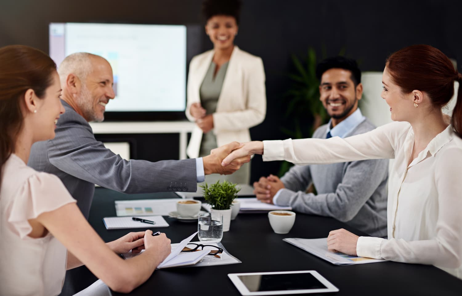 Two people shaking hands across a boardroom table after a deal