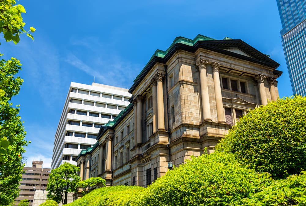 The stone head office of the Bank of Japan in Tokyo, with green copper roofs, seen from below through trees against a blue sky